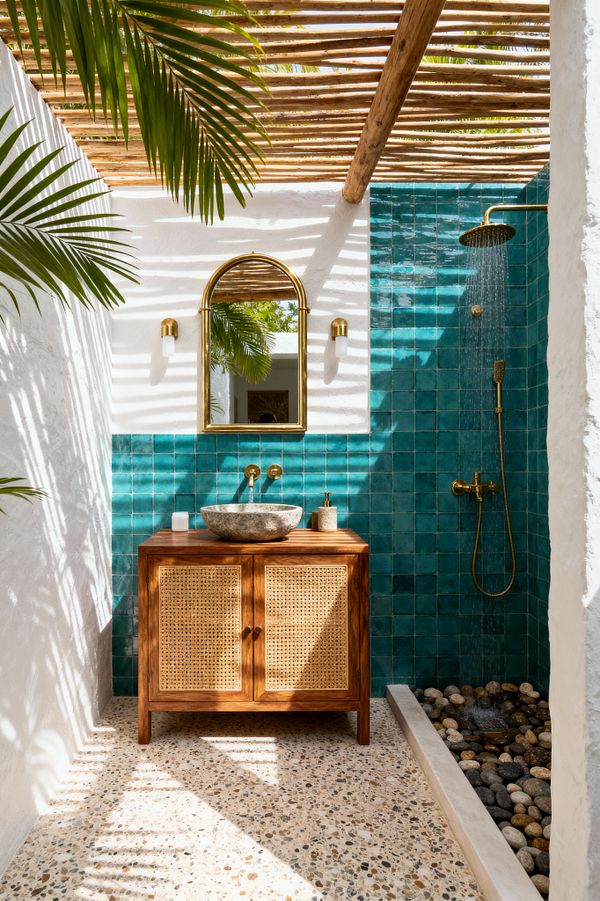 An open-air tropical bathroom with a slatted timber pergola casting striped shadows over white stucco walls. A wall of teal zellige tiles defines the vanity and shower zone, featuring a brass rain shower and hand sprayer. A warm wood cabinet with woven cane doors supports a gray stone vessel sink beneath an arched brass mirror and two small sconces. The floor is speckled terrazzo, and a shallow trough of smooth river stones collects water, with palm fronds framing the space.