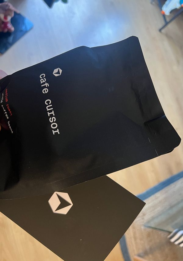 A hand holds a coffee bag and a card from Cafe Cursor.