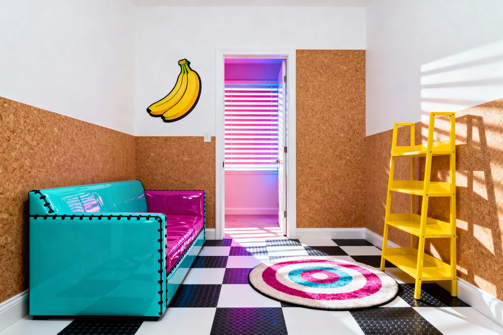 A vibrant kids’ room with half-height cork wall panels topped by white paint and a black-and-white checkerboard floor accented with textured black coin tiles. A glossy turquoise sofa with fuchsia vinyl cushions sits on the left, a bright yellow ladder-style bookcase stands on the right, and a circular target rug lies near a doorway glowing with pink and blue light through blinds, beneath cartoon banana wall art.