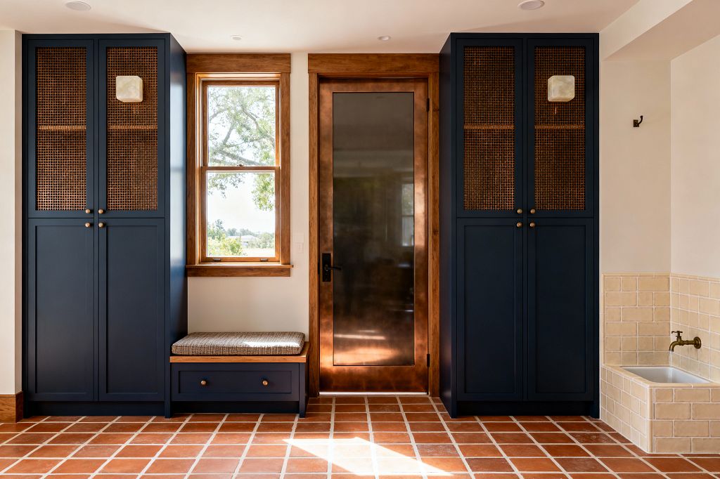 A transitional mudroom shows symmetrical navy built-in cabinets with woven cane upper doors and brass knobs flanking a wood-trimmed window and a wood-framed glass door. A cushioned bench with a drawer sits beneath the window on a grid of terracotta floor tiles with white grout. To the right, a cream-tiled utility sink with a brass wall-mounted faucet is integrated. The walls are off-white, the ceiling has recessed lights, and the door hardware is black.