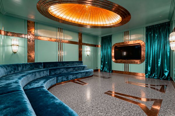 A glamorous Art Deco media room with glossy mint wall panels trimmed in patinated copper. A curved, tiered banquette in deep teal velvet wraps the space, facing a TV set into a rounded copper frame. The terrazzo floor is inlaid with copper chevrons. Overhead, an oval backlit amber glass dome is ringed in copper. Teal satin curtains and ribbed glass wall sconces complete the room’s rich, streamlined look.