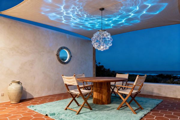 An open-air dining nook at dusk features a round wooden pedestal table with four canvas director’s chairs on a plush aqua rug over terracotta hexagon tiles. A bubble-glass chandelier hangs beneath a fabric canopy, casting watery blue reflections on the ceiling; a plastered wall with a metal-rimmed porthole window and a large ceramic jar sit to the side, with the ocean visible beyond a low parapet.