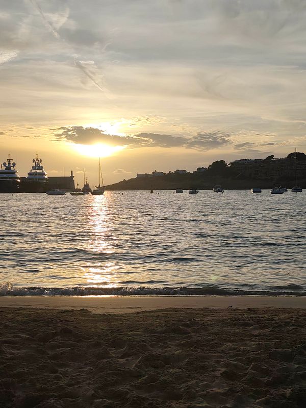 Sunset view over the sea with boats anchored near the shore and a sandy beach in the foreground.