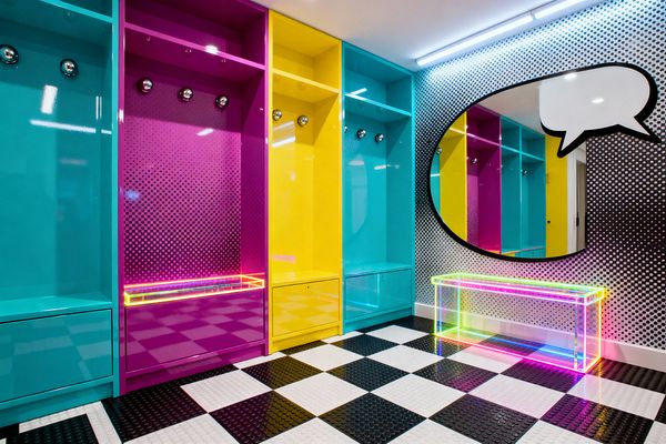 Color-blocked mudroom with glossy cyan, magenta, and yellow lockers, each with chrome hooks and bench drawers, set against walls printed with black-and-white halftone dots. A large oval mirror outlined as a comic speech bubble hangs above a clear acrylic bench that glows in neon rainbow edges. The floor is a black-and-white checkerboard with raised coin texture, while linear LED lights brighten the saturated surfaces and reflective finishes.