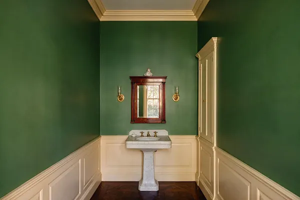 A traditional powder room with deep green walls above cream wainscoting, a dark wood framed mirror, and a white marble pedestal sink centered on the back wall. Brass wall sconces flank the mirror, while a tall built-in cabinet in a soft cream finish adds storage on the right.
