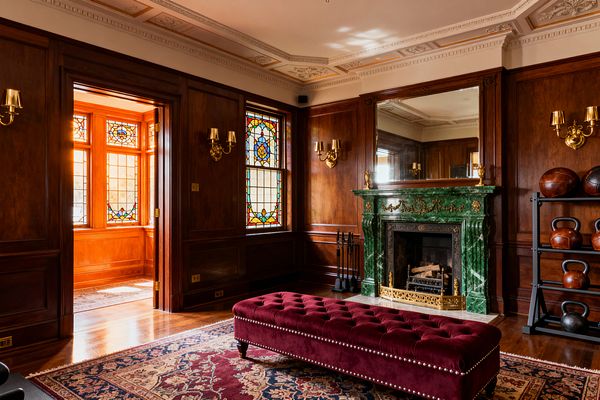 A richly paneled Victorian room functions as a home gym. A green marble fireplace with gold detailing and a brass firescreen sits at center, topped by a large mirror and flanked by brass sconces. Stained-glass windows cast warm light. A long tufted burgundy velvet bench rests on a Persian-style rug, while a black metal rack holds leather medicine balls and black kettlebells. Polished hardwood floors and ornate plaster ceiling moldings complete the space.