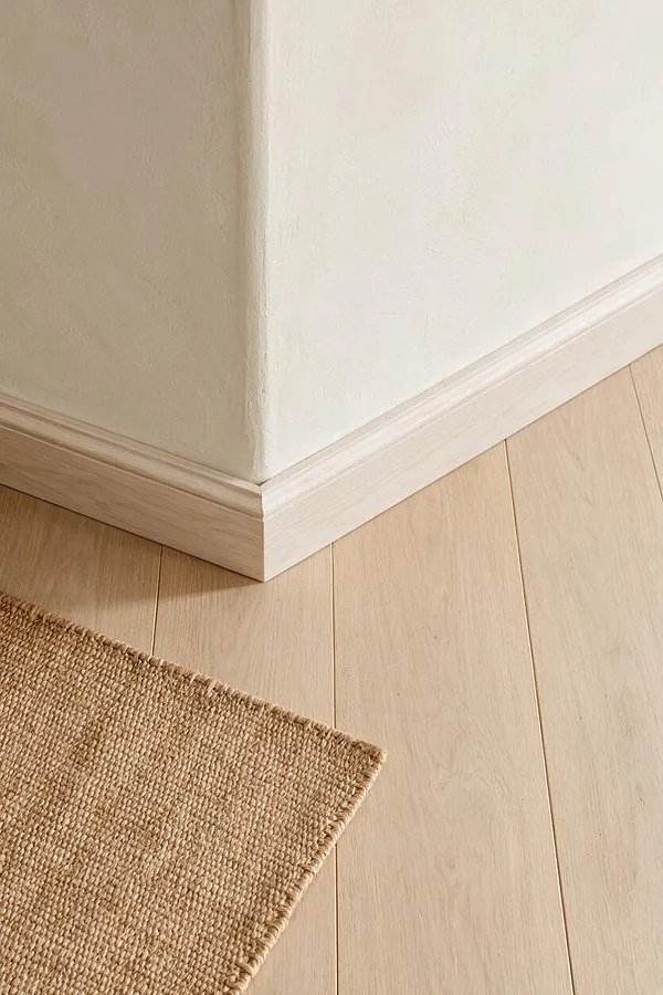 A close-up view of a Scandinavian-style interior corner showing pale wood flooring meeting soft off-white walls and matching white baseboards. A natural woven jute rug enters from the lower left, adding earthy texture to the calm neutral palette.