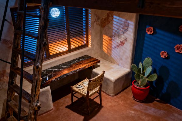 A compact Southwestern home office features adobe-textured plaster walls and a terracotta floor. A built-in bench holds a live-edge wood and black marble desk with a woven natural-fiber chair beneath wood blinds that cast warm and cool moonlit stripes. A rustic timber ladder leans nearby. A deep blue accent wall is decorated with clay rose appliqués, and a prickly pear cactus in a bright red ceramic pot anchors the corner.