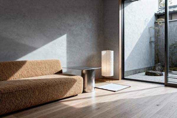 A minimalist living room with a low, camel-colored bouclé sofa set against smooth gray plaster walls. A brushed stainless-steel side table sits beside a tall cylindrical rice-paper floor lamp on a small tatami mat near a black-framed sliding glass door. Pale oak plank flooring catches sunlight that forms sharp triangular shadows. Beyond the glass is a compact Japanese courtyard with white gravel, mossy rocks, a bamboo fence, and a small tree.