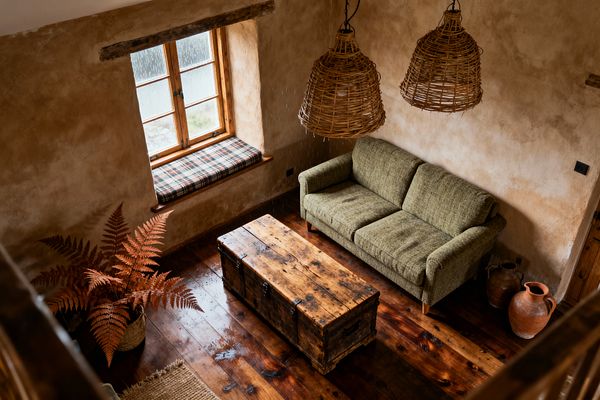 Bird’s-eye view of a rustic living room with tan plaster walls and dark polished wood floors. An olive green sofa sits against the wall beside two terracotta jugs, facing a weathered wooden trunk coffee table with black iron hardware. A wood-framed window with a plaid-cushioned sill brings light, while two large rattan pendant lights hang above; a woven basket with rust-colored ferns and a small jute rug complete the warm, earthy scene.