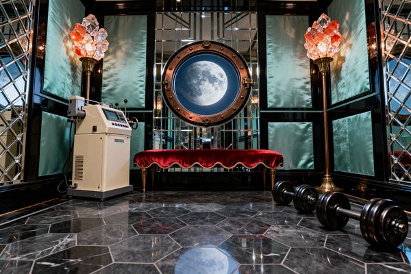 A glamorous home gym with black lacquer paneling and aqua satin insets is wrapped in diamond-beveled mirrored walls. Centered on the back wall is a round brass porthole window showing a moon image. Below sits a scalloped red velvet bench on slender gold legs, flanked by brass floor lamps topped with faceted crystal clusters. A cream exercise machine stands to the left and black dumbbells rest on dark gray marble hexagon tiles.