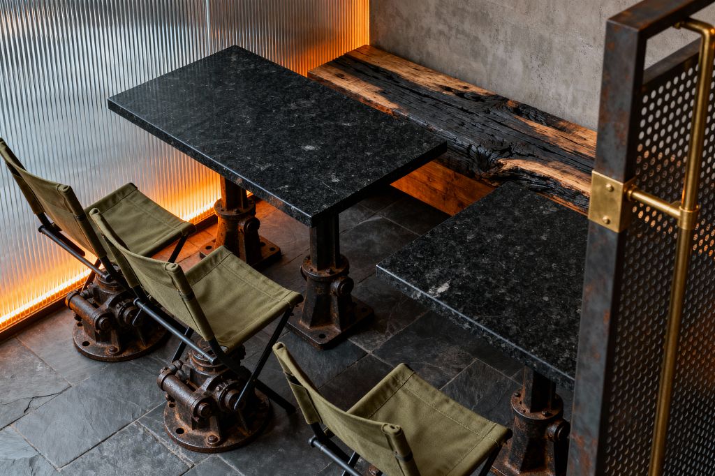 A compact industrial dining nook features two rectangular black stone tables on bolted cast-iron pedestals. Three olive green canvas sling stools sit on heavy, rusted machinery bases opposite a charred live-edge timber bench against a raw concrete wall, with a ribbed translucent panel backlit in warm amber; dark slate tiles and a perforated steel partition with a brass handle frame the scene.