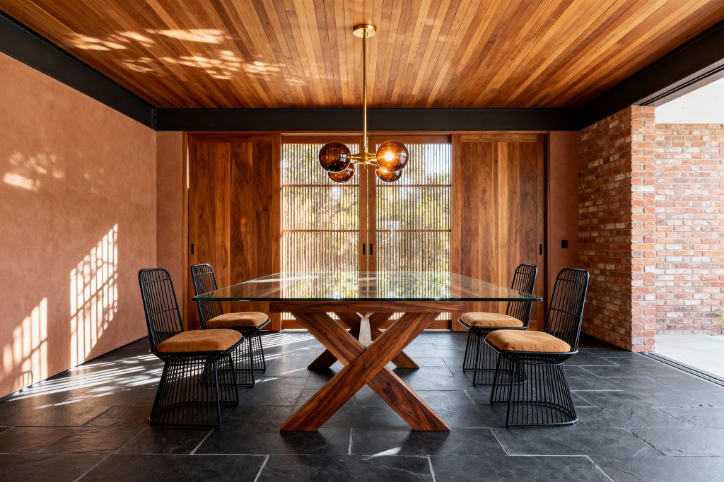 A Mid-Century Modern dining room with a rectangular glass-top table on a sculpted walnut X-base, surrounded by four black wire chairs with camel-colored cushions. The room features a warm wood slat ceiling, terracotta plaster walls, sliding walnut doors with slatted screens, a charcoal slate floor, and a brass pendant with four amber glass globes, with sunlight casting geometric shadows across the space.