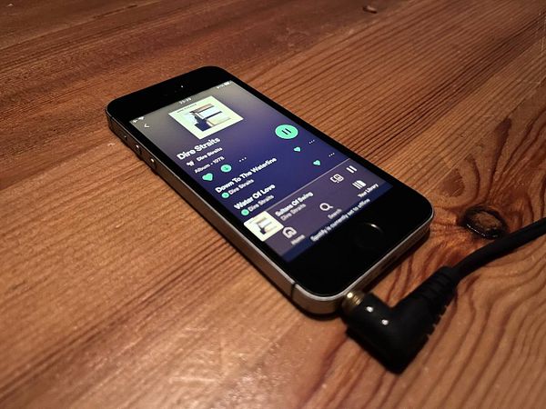 An old iPhone SE 2016 is displaying the Spotify app with a song playing.
