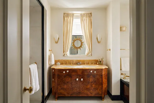 A compact master bathroom with a rich burl-wood vanity topped in warm stone, centered beneath a window with sheer curtains and a gold sunburst mirror. Brass fixtures, black trim, and cream walls create a refined Art Deco look with luxurious contrast.