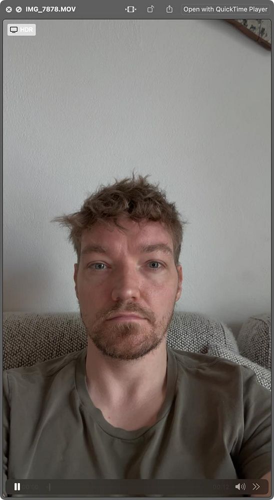 A person with tousled hair and a beard is sitting on a couch, looking directly at the camera.