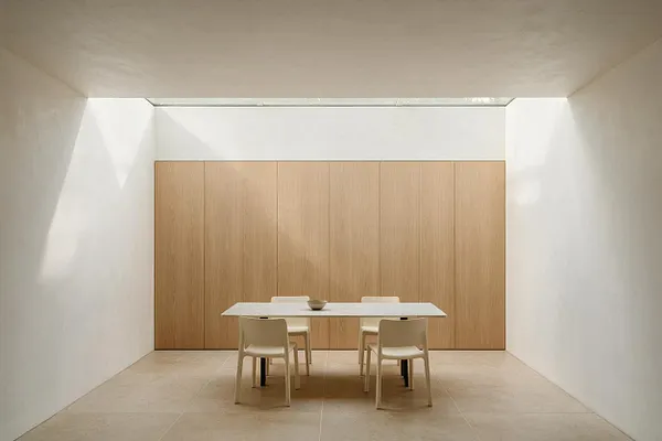 A minimalist sunroom with white walls, a pale stone tile floor, and a full back wall of light oak paneling. A simple rectangular table with four light chairs sits centered beneath a narrow skylight, creating a calm, airy space filled with soft natural light.