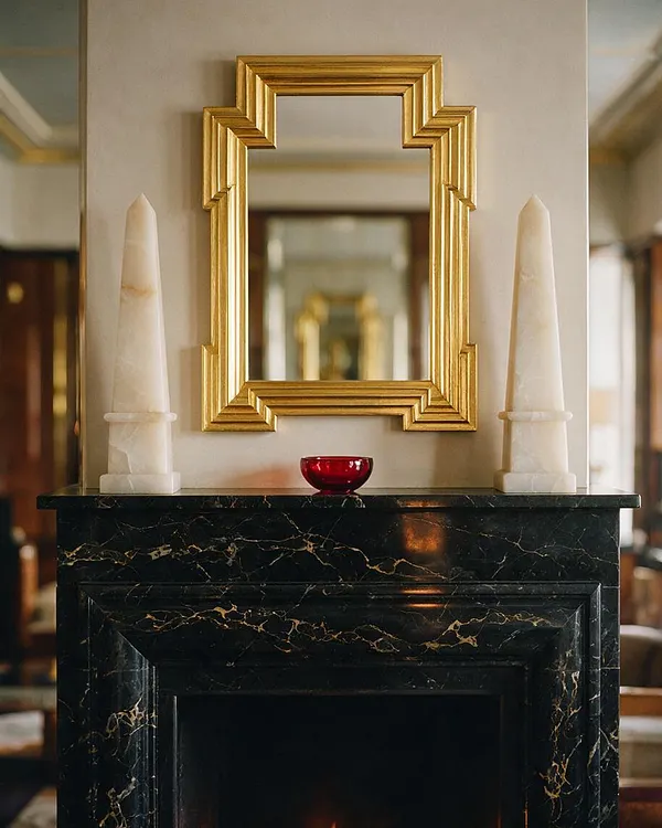 An Art Deco-inspired fireplace vignette with a black marble mantel, a geometric gold-framed mirror, and two tall ivory decorative obelisks placed symmetrically on either side. A small red bowl sits centered on the mantel, while soft neutral walls and blurred reflections in the background add warmth and depth.