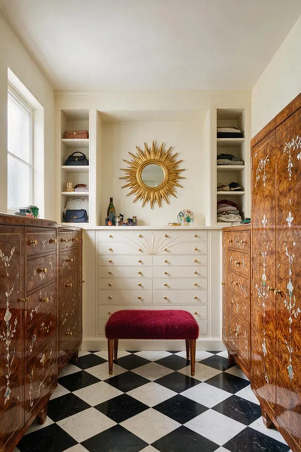 A luxurious Art Deco closet with black-and-white checkerboard flooring, glossy inlaid wood wardrobes on both sides, and a central cream built-in dresser. A gold sunburst mirror hangs above the drawers, with open shelving around it, and a burgundy upholstered bench sits in the foreground.