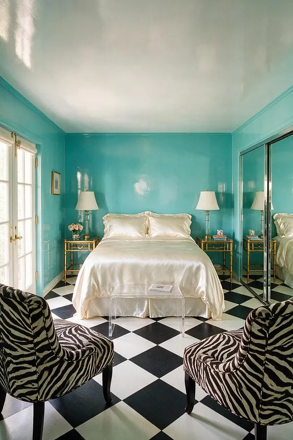 A glamorous guest bedroom with glossy turquoise walls, a white satin bed, gold nightstands, and mirrored closet doors. Black-and-white checkered flooring and zebra-patterned accent chairs add dramatic Hollywood Regency style.