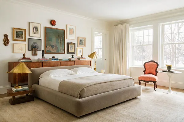 A bright eclectic master bedroom with a low upholstered bed, layered neutral bedding, and a wood credenza topped with books and sculptural objects. The wall above is filled with a curated mix of framed artwork, and the room is accented by brass lamps, an orange vintage chair, and large windows with cream curtains.