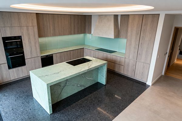 A contemporary U-shaped kitchen with a mint green stone waterfall island holding an induction cooktop. Flat-panel light oak cabinets span two walls with matching stone countertops and a pale aqua back-painted glass backsplash. Black built-in double ovens and a concealed dishwasher sit flush in the cabinetry. A light-gray plaster hood anchors a second induction zone. Dark terrazzo flooring defines the work area beside lighter tile, under a circular cove of warm recessed lighting.