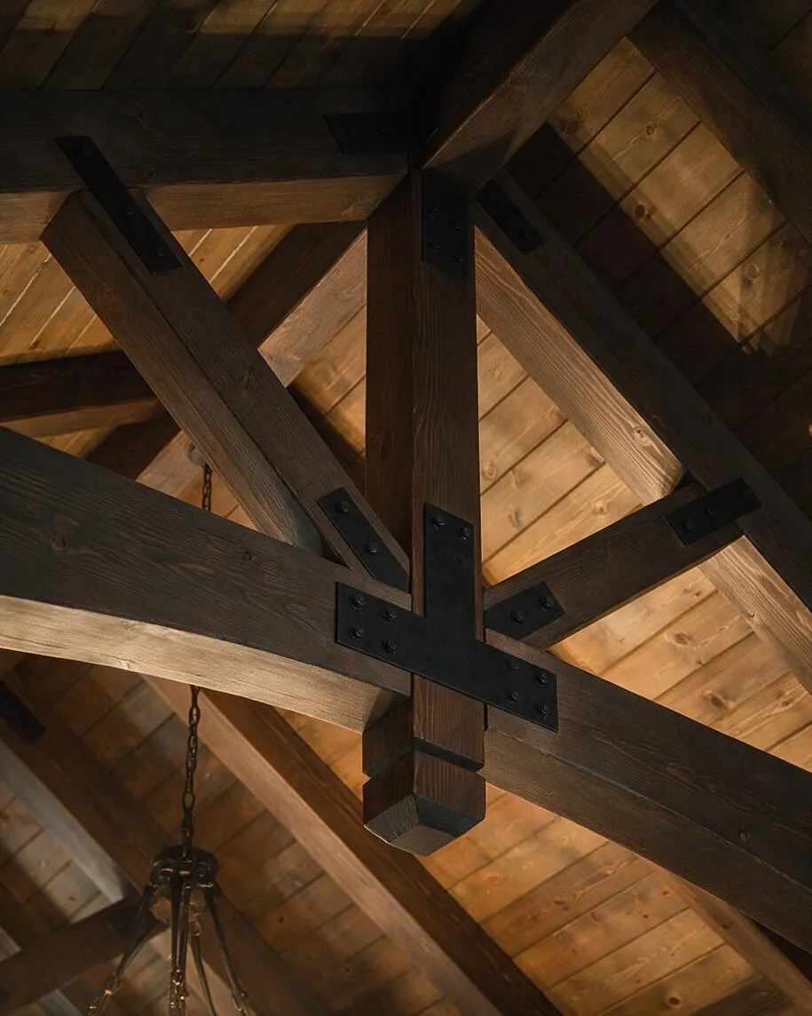 A close-up view of rustic ceiling construction with intersecting heavy wood beams and diagonal braces, joined by black metal plates and bolts. Warm natural wood planks fill the background, creating a cozy, lodge-like atmosphere.