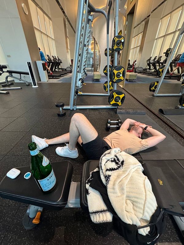 A person is resting on the gym floor next to a bench and weights.
