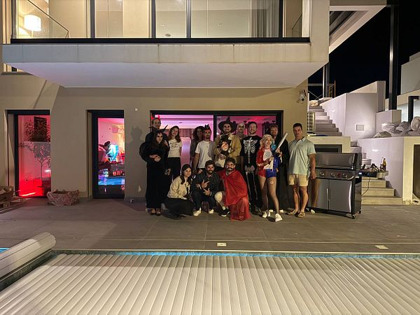 A group of friends dressed in Halloween costumes poses in front of a modern house during a BBQ party.