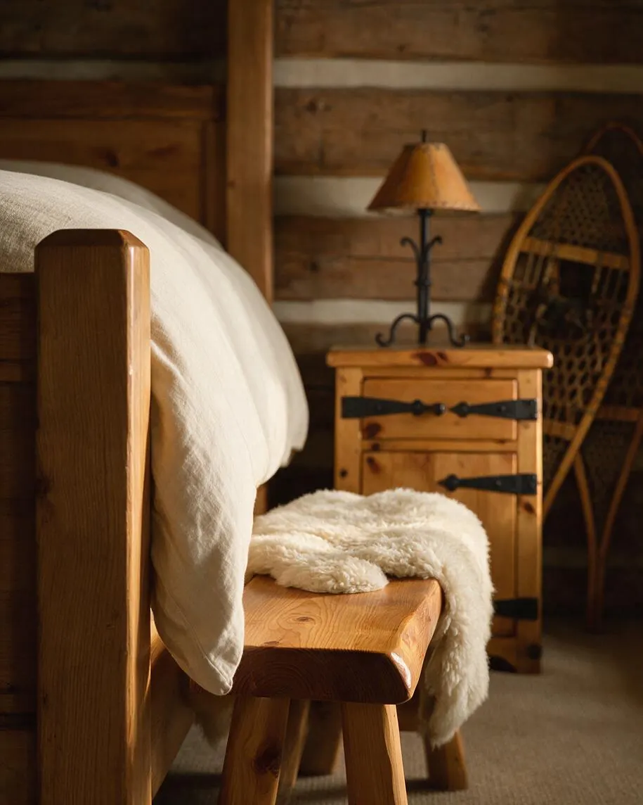 A rustic bedroom vignette with a wooden bed frame, cream bedding, and a small matching bench topped with a sheepskin throw. A wooden nightstand with black iron hardware and a warm-toned lamp sits beside the bed, while a woven snowshoe-like wall piece adds texture against the log wall.