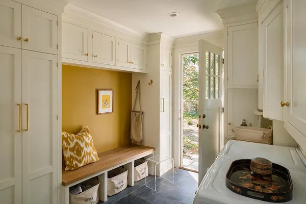 A bright mudroom with floor-to-ceiling white built-in cabinets, brass hardware, and a wood bench set into a mustard-yellow recessed nook. Open cubbies hold fabric storage bins beneath the bench, while a patterned pillow and woven tote add texture. A glass-paneled door brings in natural light beside a utility sink and laundry equipment.