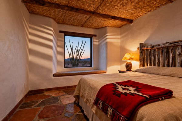 A cozy Southwestern bedroom features smooth plaster walls and a woven reed ceiling with exposed timber vigas. A rustic log headboard anchors a bed in sand linen with a red geometric wool throw, while terracotta flagstone floors, a warm stone table lamp, and a deep square window with cushioned sill frame a desert sunset that casts striped shadows across the walls.