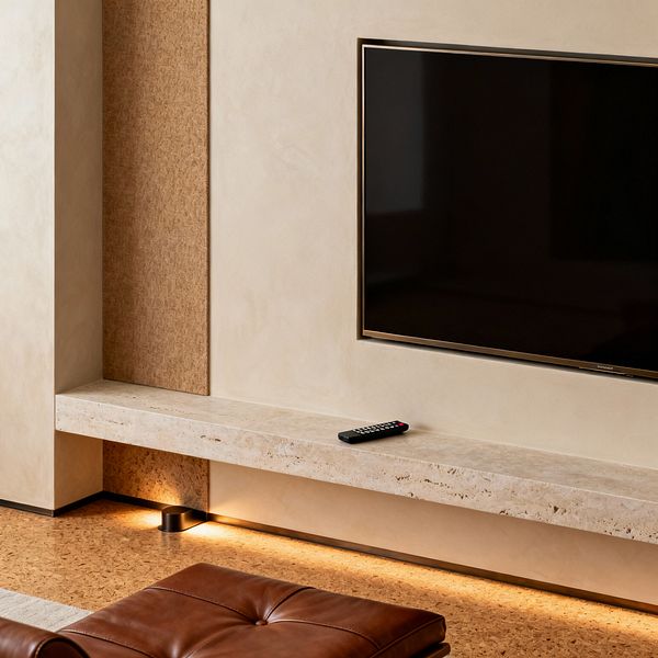Close-up of a minimalist media room showing a wall-mounted flat-screen TV with a slim dark frame above a floating travertine shelf. Smooth warm-beige plaster walls and a narrow tan textured panel provide subtle contrast, while concealed LED lighting beneath the ledge casts a soft amber glow across natural cork flooring. A tufted caramel leather ottoman is visible in the foreground, and a remote control rests on the stone shelf.