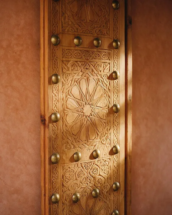 A close-up of a richly carved wooden door panel framed by warm terracotta plaster walls. The panel features intricate geometric and floral Moroccan-style carvings, accented with rows of round brass studs that catch the light.