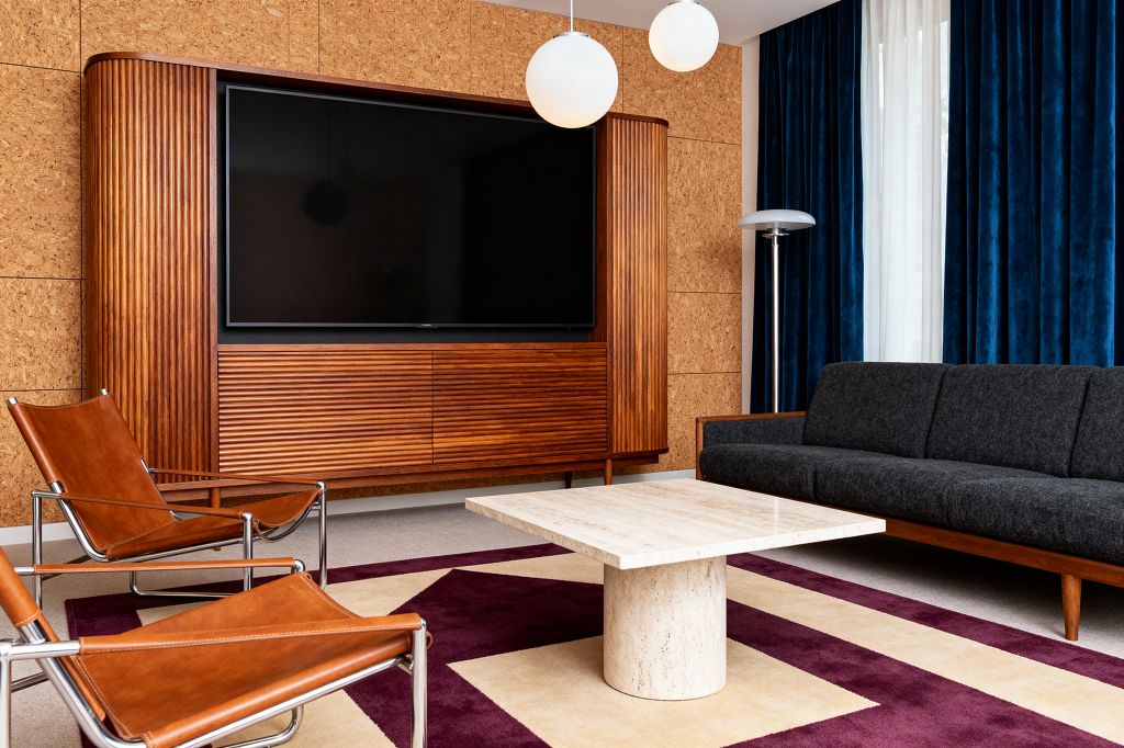 A Mid-Century Modern media room features a large TV set into a walnut fluted cabinet against cork wall panels, a dark charcoal sofa with wood base, and two cognac leather sling chairs on chrome frames. A square travertine pedestal coffee table sits on a cream-and-plum geometric rug, with deep blue velvet drapes and white sheers, twin globe pendant lights, and a silver mushroom floor lamp completing the cozy, acoustically softened space.