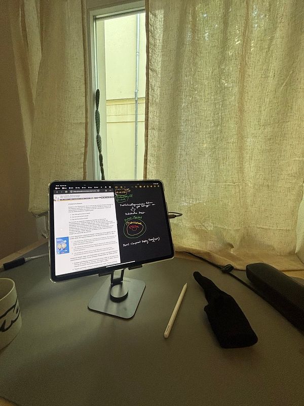 A workspace featuring an iPad on a stand, a coffee mug, and a pen, with natural light filtering through sheer curtains.