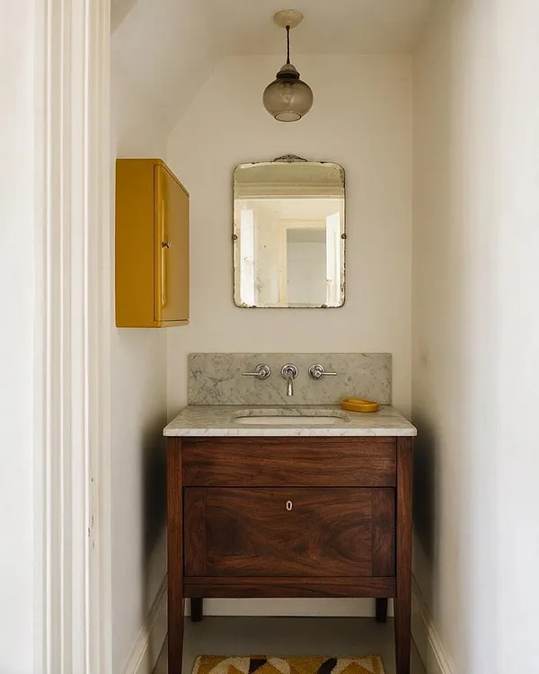 A narrow eclectic master bathroom with a dark walnut vanity topped by white marble, a simple wall-mounted sink faucet, and a small aged mirror above it. A mustard yellow wall cabinet adds a bold accent against the white walls, while a single globe pendant light hangs from the ceiling.