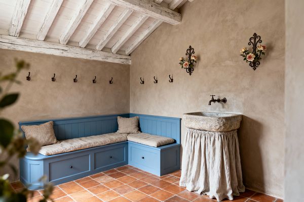 A rustic French Country mudroom featuring an L-shaped built-in blue bench with paneled fronts and storage drawers, topped with beige linen cushions. Hand-troweled beige plaster walls display a row of wrought-iron hooks and two floral iron plaques. A rough-hewn stone trough sink with a gathered linen skirt and a wall-mounted bronze faucet stands over terracotta floor tiles beneath whitewashed exposed ceiling beams.