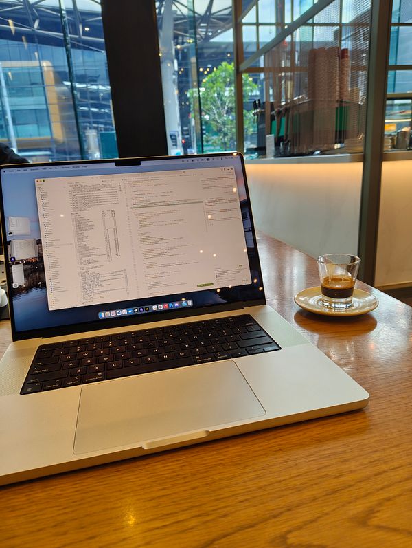 A laptop is open on a wooden table in a cafe, displaying code, alongside a small espresso on a saucer.