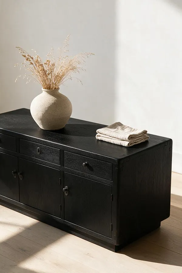 A low black wooden sideboard stands against a light wall on a pale wood floor. A textured cream vase with dried grasses sits on top beside a folded neutral linen cloth, creating a calm Japandi-inspired vignette.