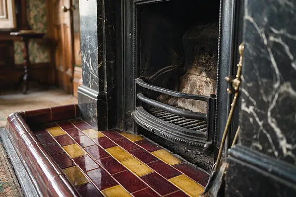 A close-up of a Victorian fireplace featuring a black cast-iron firebox surround with brass andirons and a glossy tiled hearth. The hearth is made of burgundy and mustard-yellow rectangular tiles set in a decorative pattern, framed by dark marble with white veining.