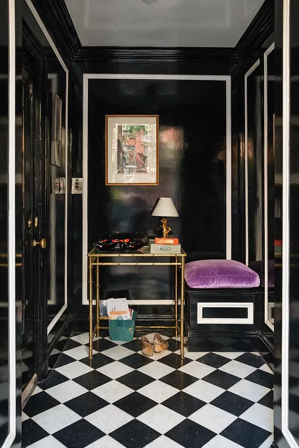A narrow Hollywood Regency entryway with glossy black walls outlined in white trim, a black-and-white diamond checkerboard floor, and mirrored panel details. A slim brass console table holds a small lamp and decorative bowl, while a purple velvet cushion sits on a black storage bench beside it.