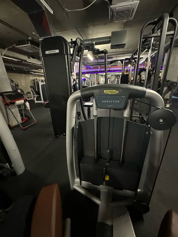 The image captures a gym interior featuring a Technogym adductor machine.