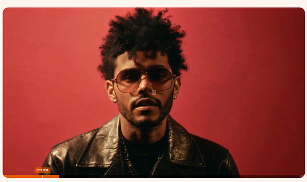 A video player interface featuring a man with curly hair and sunglasses against a red background.