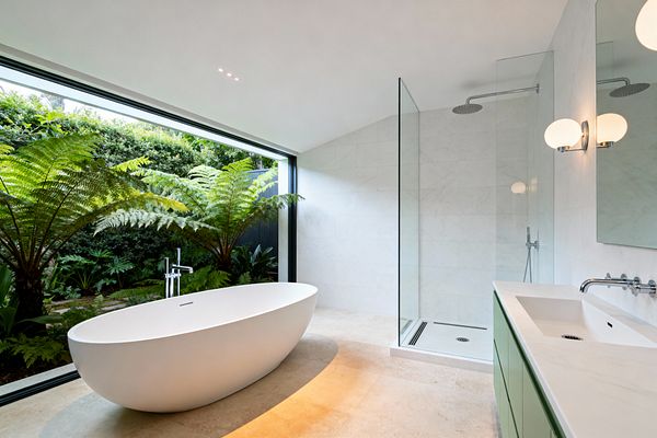 A modern bathroom features a floor-to-ceiling glass wall overlooking a lush fern garden. Inside, a matte white oval freestanding tub sits on warm beige stone flooring beside a frameless walk-in shower with a ceiling-mounted rain head. A vanity with sage green flat-panel doors and a white countertop includes a wall-mounted faucet below a wide mirror and a globe sconce; black window frames and chrome fixtures complete the minimal look.