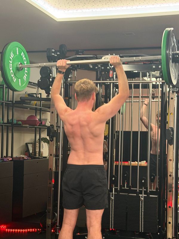 A shirtless man in black shorts performing an overhead barbell press in a gym.