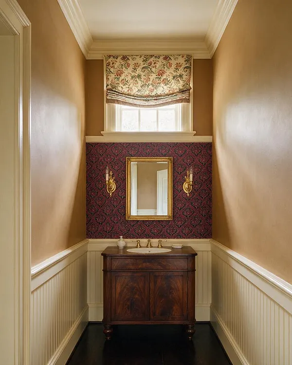 A narrow traditional powder room with warm tan walls, white wainscoting, and a dark wood vanity centered beneath a gold-framed mirror. Burgundy floral wallpaper and a patterned Roman shade add rich texture above the sink, while brass sconces and polished fixtures create a classic, formal look.