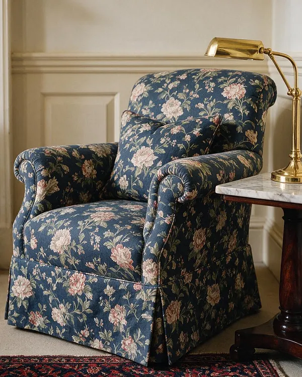 A traditional upholstered armchair in dark blue floral fabric sits beside a marble-topped side table with a polished brass desk lamp. The chair has rolled arms, a plush cushion, and a skirted base, set against cream paneled walls and a patterned rug.