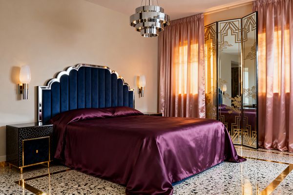 A glamorous Art Deco bedroom with a scalloped, channel-tufted navy velvet headboard trimmed in chrome behind a bed dressed in plum satin. Black textured nightstands with brass bases sit on each side under opal glass sconces. A tiered chrome chandelier hangs overhead. To the right, blush pink curtains frame a mirrored screen etched with gold geometric and floral motifs. The room has cream walls and a terrazzo floor with brass inlay, reflecting warm evening light.