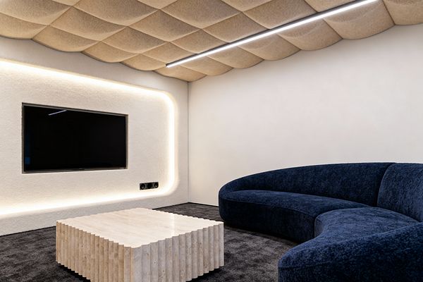 A contemporary media room with a deep navy curved sofa on a charcoal carpet facing a wall-mounted TV recessed into a rounded niche. Perimeter LED lighting traces the niche, and black circular speaker grilles sit below the screen. The ceiling is clad in beige, quilted felt acoustic panels with a slim linear LED light. Walls are smooth warm white plaster, and a low, fluted travertine coffee table anchors the space, creating a quiet, minimal, theater-like setting.